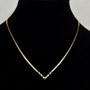 May include: A gold-toned necklace with a V-shaped pendant. The pendant is adorned with alternating green and clear gemstones. The necklace is displayed against a black background, highlighting the jewelry's design and sparkle.