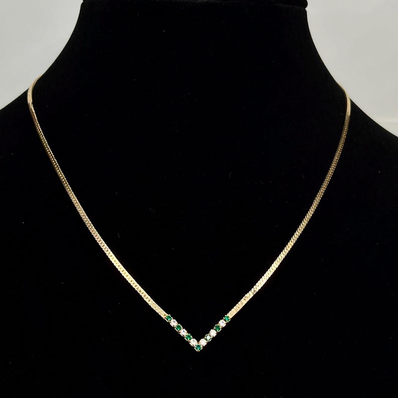 May include: A gold-toned necklace with a V-shaped pendant. The pendant is adorned with alternating green and clear gemstones. The necklace is displayed against a black background, highlighting the jewelry's design and sparkle.