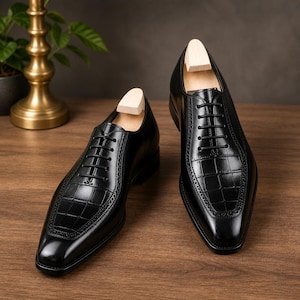 May include: A pair of black leather dress shoes with a crocodile-textured vamp and toe. The shoes have a classic lace-up design and are displayed on a wooden surface. Wooden shoe trees are inserted inside the shoes.