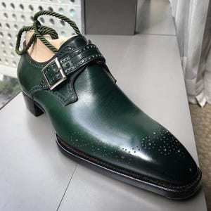 May include: A single, dark green leather shoe with a buckle closure and decorative perforations. The shoe has a pointed toe and a small heel. A green rope is attached to the shoe for display.