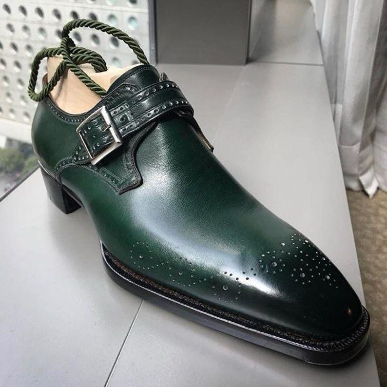 May include: A single, dark green leather shoe with a buckle closure and decorative perforations. The shoe has a pointed toe and a small heel. A green rope is attached to the shoe for display.