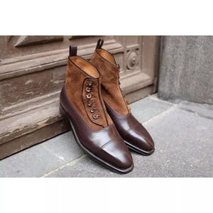 Puede incluir: Botas de cuero marrón con parte superior de ante y cierres de botón. Las botas tienen una punta puntiaguda y un tacón bajo.