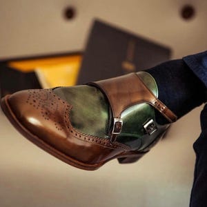 Könnte beinhalten: Nahaufnahme eines braunen und grünen Leder-Doppelmonk-Schuhs. Der Schuh hat ein Flügelspitzendesign mit dekorativen Perforationen und einem Schnallenverschluss. Der Schuh ist ein klassischer Stil.
