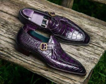 May include: A pair of purple leather dress shoes with a crocodile pattern, featuring a buckle and tassel detail. The shoes are displayed on a wooden bench, showcasing their polished finish and elegant design.
