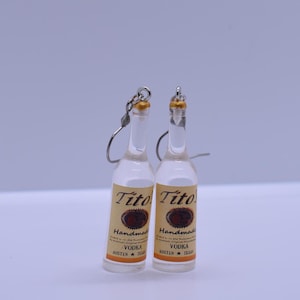 May include: A pair of silver dangle earrings featuring miniature clear glass bottles with gold tops. The bottles have a label that reads "Tito's Handmade Vodka" with the words "Austin, Texas" at the bottom.