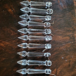 May include: A collection of clear glass chandelier prisms with pointed ends and decorative faceted details. Each prism has a small wire loop for hanging. The prisms are arranged on a dark wooden surface.