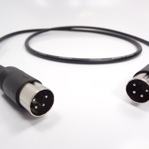 Puede incluir: Un cable MIDI negro con dos conectores DIN de 5 pines. El cable está enrollado y descansa sobre una superficie blanca. Los conectores tienen una carcasa metálica plateada y una carcasa de plástico negro.