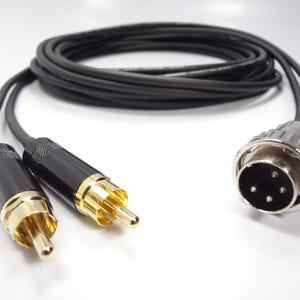 May include: A black audio cable featuring two gold-plated RCA male connectors on one end and a single four-pin XLR-style circular connector on the other. The cable is coiled in the background, showing a smooth black jacket and durable construction.