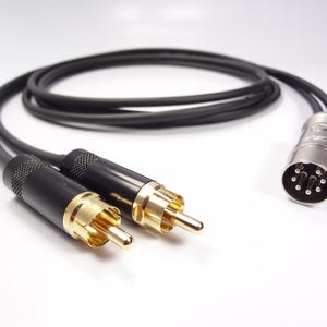 Puede incluir: Cable de audio negro con conectores RCA dorados y un conector DIN plateado de 7 pines. El cable está enrollado y descansa sobre una superficie blanca. Aproximadamente 100 cm de largo.