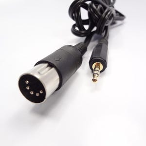 Puede incluir: Un cable de audio negro con un conector DIN de cinco pines en un extremo y un conector estéreo chapado en oro de 3,5 mm en el otro. El cable está enrollado y la carcasa de plástico negro del conector DIN muestra una pequeña flecha.