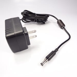 Op de afbeelding: Een zwarte AC-adapter met een tweepolige stekker en een zwarte kabel. De kabel is opgerold bij de adapter en loopt door naar een DC-connector. De adapter heeft een rechthoekige vorm met drie verticale lijnen aan één kant.