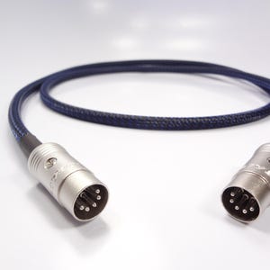 Puede incluir: Un cable de audio trenzado azul y negro con conectores plateados en cada extremo. Los conectores tienen múltiples pines y son de forma cilíndrica. El cable está curvado y descansa sobre una superficie blanca.