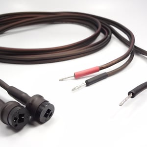Peut inclure: Une paire de câbles audio marron enroulés en cercle. Chaque câble présente un connecteur circulaire noir à une extrémité et deux fils conducteurs exposés à l'autre. Les fils sont terminés par une gaine thermorétractable rouge et noire.