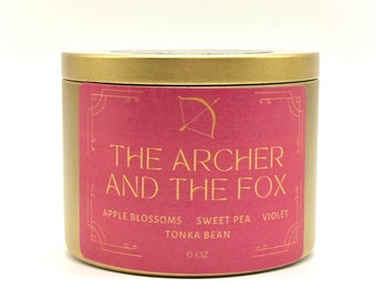 May include: Gold candle tin with a pink label that reads "The Archer and the Fox" with a gold arrow and bow graphic. The label also lists the scent notes: Apple Blossoms, Sweet Pea, Tonka Bean, and Violet. The candle is 6 oz.