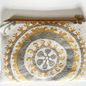 Op de afbeelding: Een rechthoekig etui met een gouden rits. Het ontwerp heeft concentrische cirkels in grijs en goud op een crèmekleurige achtergrond. Een klein, bruin leren label is aan de zijkant bevestigd.