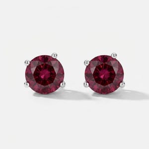May include: A pair of stud earrings featuring round-cut, deep red gemstones set in silver-toned metal. Each stone is secured by three small prongs, creating a circular silhouette that highlights the vibrant, saturated color of the faceted gems.