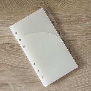 May include: A translucent white plastic planner divider with a curved pocket on the front and six circular holes along the left edge. The divider features several protruding tabs along the right side, each labeled with text including Groceries, Gas, Spending, Gifts, Eat out, and Misc.