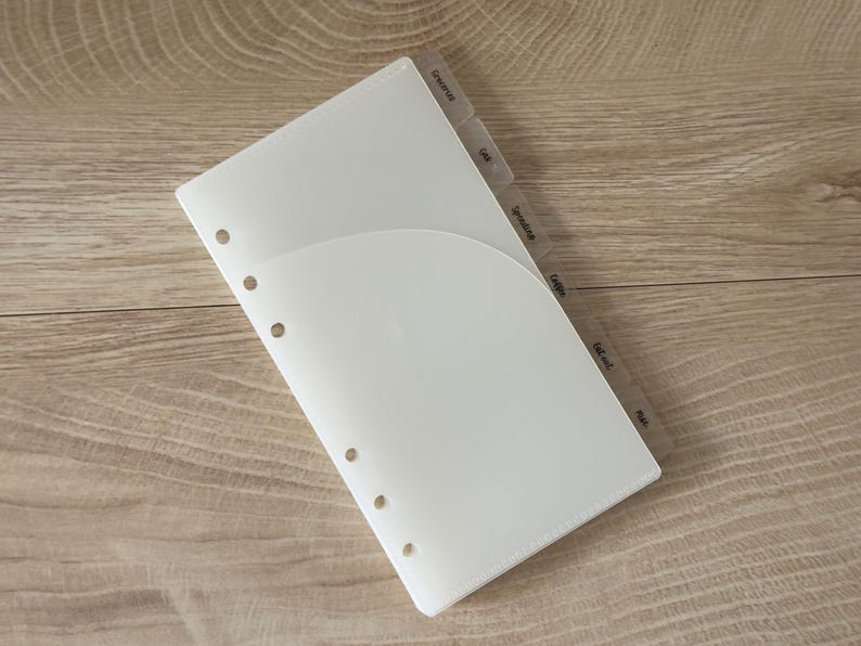 May include: A translucent white plastic planner divider with a curved pocket on the front and six circular holes along the left edge. The divider features several protruding tabs along the right side, each labeled with text including Groceries, Gas, Spending, Gifts, Eat out, and Misc.