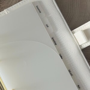 May include: A white textured binder featuring gold-toned metal rings and translucent plastic dividers. The dividers are labeled with black text reading Groceries, Gas, Spending, Coffee, Eat out, and Misc. The binder has a pen loop on the side.