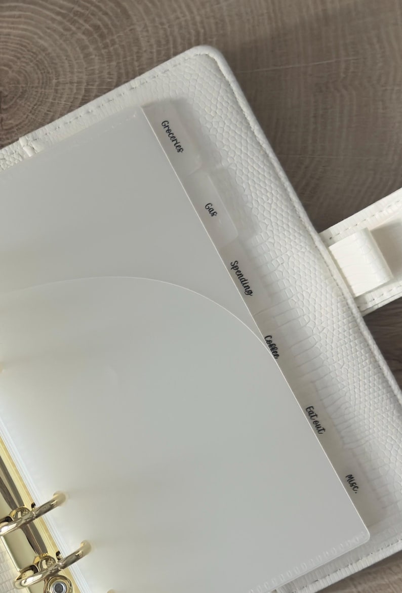 May include: A white textured binder featuring gold-toned metal rings and translucent plastic dividers. The dividers are labeled with black text reading Groceries, Gas, Spending, Coffee, Eat out, and Misc. The binder has a pen loop on the side.