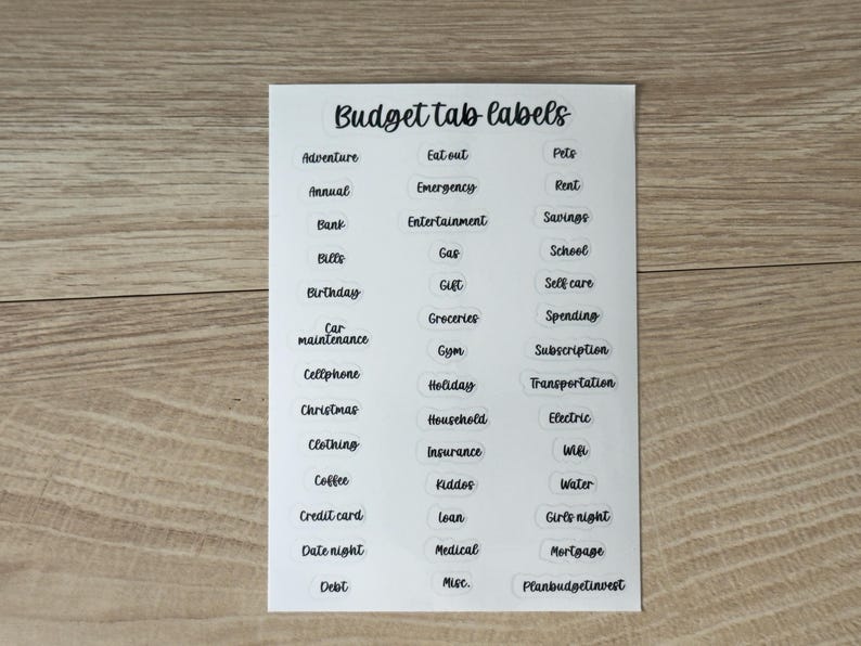 May include: A white sticker sheet titled Budget tab labels in black cursive font. It contains three columns of individual transparent labels with black text for various categories including Adventure, Rent, Savings, and Debt, organized for organization.