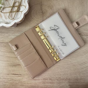 May include: A beige planner with gold accents, open to reveal a clear pocket labeled "Spending." The planner has a textured exterior, multiple card slots, and a gold binder mechanism. The planner is on a light wood surface.