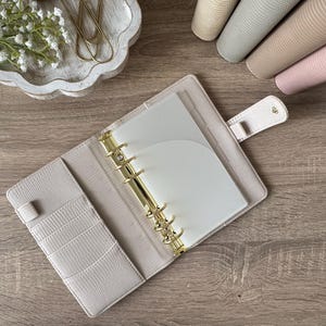 May include: An open beige planner with a textured cover, featuring a gold six-ring metal binder mechanism, multiple interior card slots, and a snap-button closure tab. The planner contains white paper inserts and a translucent plastic divider.