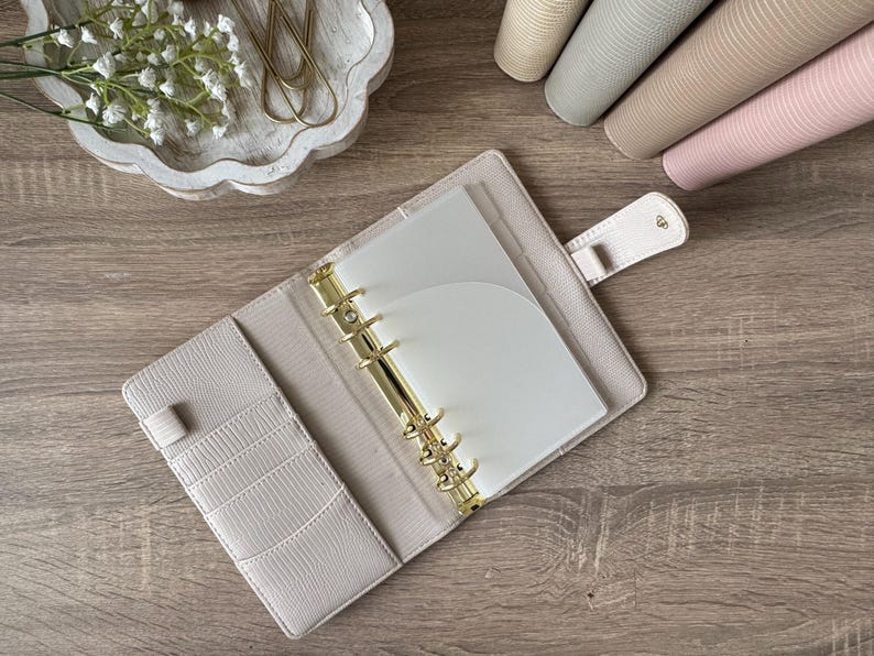 May include: An open beige planner with a textured cover, featuring a gold six-ring metal binder mechanism, multiple interior card slots, and a snap-button closure tab. The planner contains white paper inserts and a translucent plastic divider.