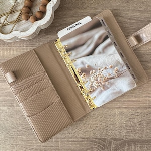 Pu&ograve; includere: Agenda beige aperta con anelli dorati e divisori con le etichette "Spending", "Beauty", "Nails", "Crafts", "Barber" e "Meller". L'agenda &egrave; aperta su una pagina con un'immagine floreale e la parola "Personal". Su una superficie di legno.