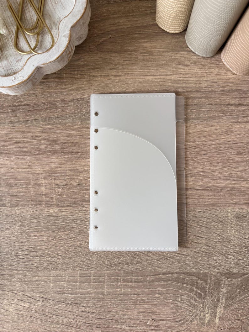 May include: A translucent, frosted plastic planner divider featuring a curved pocket on the front and six circular punched holes along the left edge. The divider includes a series of six clear tabs extending from the right side for organization.