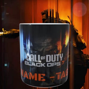 Può includere: Una tazza cilindrica in ceramica con una grafica scura ad alto contrasto di Call of Duty Black Ops 7. Il design include il volto di un soldato in ombra, il titolo del gioco in bianco e la scritta arancione NAME - TAG sul fondo della tazza.