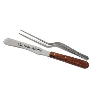 Könnte beinhalten: Ein Edelstahlspatel mit Holzgriff und dem Text "Custom Name" auf der Klinge, zusammen mit einer Edelstahlpinzette. Beide Werkzeuge sind für Präzisionsarbeiten konzipiert.