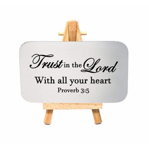 May include: A rectangular silver-toned metal plaque with rounded corners, resting on a small wooden easel. The plaque features black text that reads: Trust in the Lord With all your heart Proverb 3:5. The design is simple and functional.