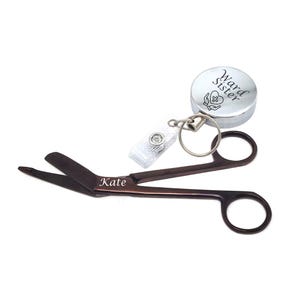 May include: Brown medical scissors with the name "Kate" engraved on the blade, next to a silver retractable badge holder with the words "Ward Sister" and a medical symbol. The scissors are open, and the badge holder is attached to a white clip.