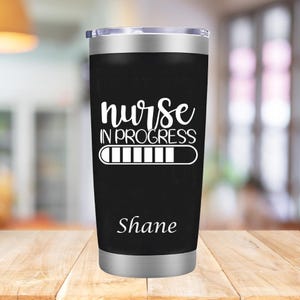 Puede incluir: Un vaso de acero inoxidable negro con un diseño blanco que dice "nurse in progress" con una barra de progreso debajo. El nombre "Shane" está impreso en la parte inferior del vaso.