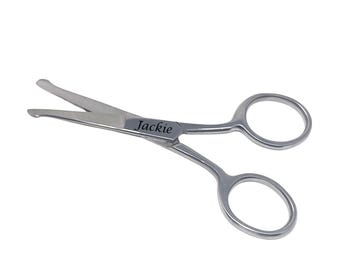 May include: A pair of small, silver-colored scissors with rounded finger holes. The scissors have a curved blade and the name "Jackie" is engraved on the handle. The scissors are designed for trimming.