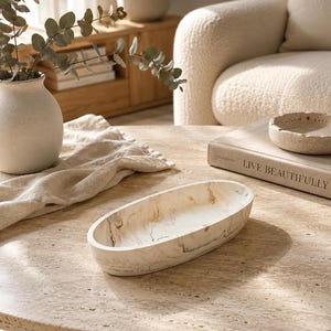 May include: An oval-shaped decorative bowl featuring a cream and beige marbled pattern with dark brown veining. The bowl has a smooth, curved interior and exterior surface, showcasing a natural stone-like appearance with a polished finish.