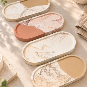 May include: Four oval-shaped trays with raised edges, each featuring a split design. One half of each tray displays a solid matte colour in tan, terracotta, cream, or beige, while the other half features a swirling marbled pattern in matching tones.