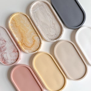 Könnte beinhalten: Satz von acht ovalen Tabletts in verschiedenen Farben, darunter rosa, beige, gelb, weiß, grau und braun. Die Tabletts haben ein marmoriertes Design und sind perfekt zum Organisieren kleiner Gegenstände.