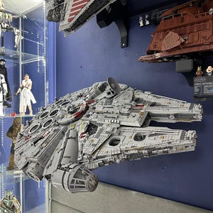 May include: A large, gray and white Lego Millennium Falcon spaceship model mounted on a wall. The model is detailed with red and yellow accents. The spaceship is hanging above a gray couch.