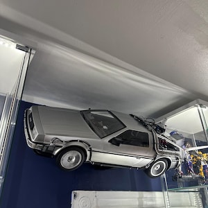 Puede incluir: Un modelo de coche DeLorean plateado, una máquina del tiempo, está montado en una pared, con el coche mirando hacia el lado izquierdo de la imagen. El coche está suspendido en el aire, como si estuviera volando. El coche está en una habitación con una pared azul y un techo blanco.