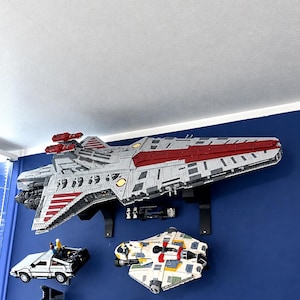 K&ouml;nnte beinhalten: Ein gro&szlig;es, grau-rotes Raumschiffmodell an einer blauen Wand, platziert &uuml;ber einem wei&szlig;en Sportwagenmodell, einem bunten Raumschiff, einem dunkelgrauen J&auml;ger und einem orangefarbenen Landspeeder, alle als Wandmodelle aus Bausteinen montiert.