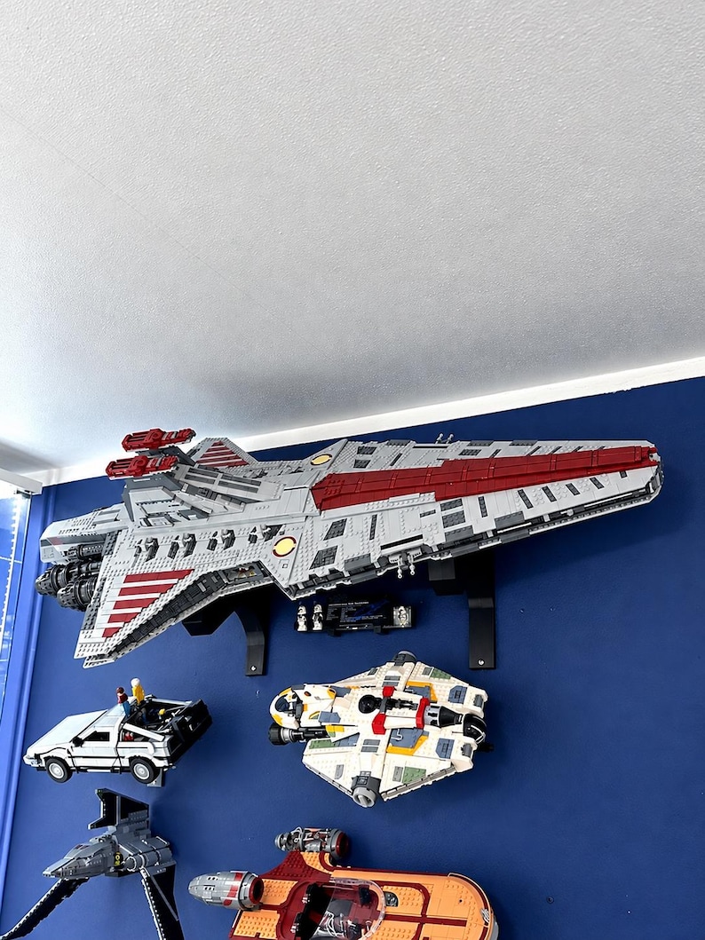 K&ouml;nnte beinhalten: Ein gro&szlig;es, grau-rotes Raumschiffmodell an einer blauen Wand, platziert &uuml;ber einem wei&szlig;en Sportwagenmodell, einem bunten Raumschiff, einem dunkelgrauen J&auml;ger und einem orangefarbenen Landspeeder, alle als Wandmodelle aus Bausteinen montiert.