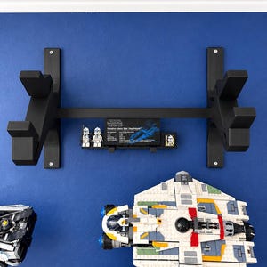 K&ouml;nnte beinhalten: Ein schwarzes Wandregal mit zwei vertikalen St&uuml;tzen und einer horizontalen Querstange. Eine kleine schwarze Plakette mit dem Text Star Wars Venator-class Star Destroyer und zwei kleine wei&szlig;-blaue Minifiguren sind an der Mittelstange befestigt.