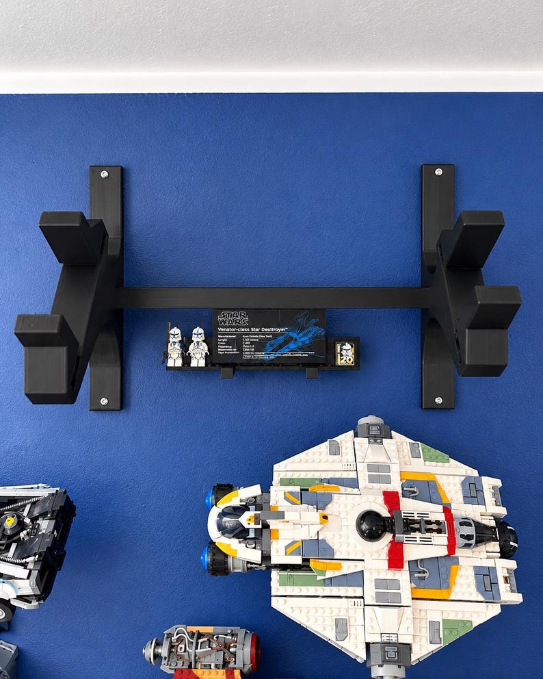 K&ouml;nnte beinhalten: Ein schwarzes Wandregal mit zwei vertikalen St&uuml;tzen und einer horizontalen Querstange. Eine kleine schwarze Plakette mit dem Text Star Wars Venator-class Star Destroyer und zwei kleine wei&szlig;-blaue Minifiguren sind an der Mittelstange befestigt.