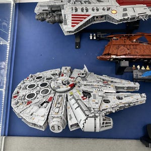 Pu&ograve; includere: Un grande e dettagliato modello di astronave LEGO grigio e rosso che ricorda il Millennium Falcon, caratterizzato da uno scafo circolare, una cabina di pilotaggio e complessi pannelli di superficie, montato su una parete blu sopra un veicolo di trasporto LEGO marrone.