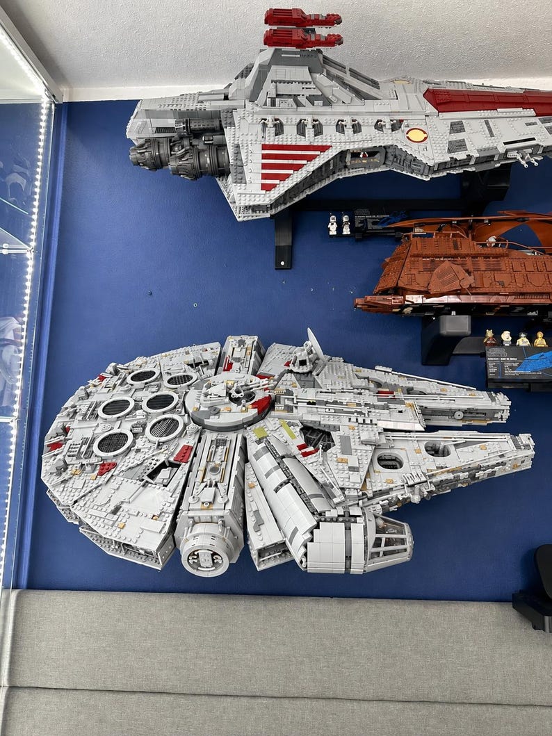 Pu&ograve; includere: Un grande e dettagliato modello di astronave LEGO grigio e rosso che ricorda il Millennium Falcon, caratterizzato da uno scafo circolare, una cabina di pilotaggio e complessi pannelli di superficie, montato su una parete blu sopra un veicolo di trasporto LEGO marrone.