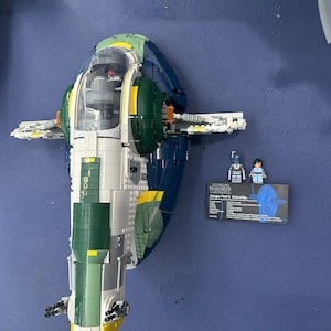 Puede incluir: Un modelo Lego de la nave espacial de Jango Fett, con detalles verdes, azules, blancos y amarillos. El modelo está montado en una pared azul. También se ve una pequeña tarjeta informativa y dos figuras de Lego.