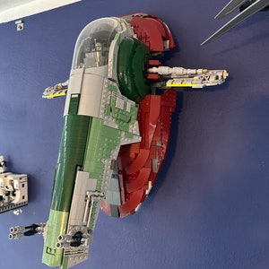 May include: A green and red Lego model of the Star Wars spacecraft Slave I, mounted on a wall. The model is detailed with a cockpit, wings, and a rear engine.