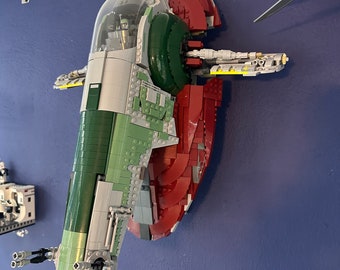 May include: A green and red Lego model of the Star Wars spacecraft Slave I, mounted on a wall. The model is detailed with a cockpit, wings, and a rear engine.